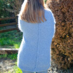Une personne debout à l'extérieur, vêtue d'une confortable Veste en mohair de chevreau Vannerie tricotée à la main bleue, dos à la caméra et la lumière du soleil filtrant à travers les arbres en arrière-plan.