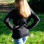 Une personne aux longs cheveux blonds, debout dans un jardin, les mains sur les hanches, portant un pull Pull Mousse col boule taille unique en kid-mohair et soie naturelle et un jean bleu, face à la caméra.