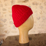 Une tête de mannequin en bois affichant un bonnet tricoté rouge vif, en mohair éthique, sur un fond de mur en pierre rustique.