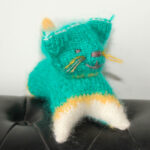 Un jouet pour chat tricoté à la main en turquoise avec un détail orange et blanc repose sur une surface noire, mettant en valeur ses charmantes moustaches et son expression borgne espiègle. Cet article unique est fabriqué à partir de