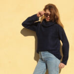 Jeune femme en Pull Mousse col boule taille unique en kid-mohair et soie naturelle bleu marine tendance et un jean classique posant sur un fond jaune chaud, jetant un coup d'œil ludique par-dessus ses lunettes de soleil.
