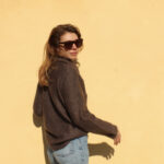 Une femme portant des lunettes de soleil et un confortable Pull Mousse col boule taille unique en kid-mohair et soie naturelle debout contre un mur jaune vif, tournant la tête pour regarder par-dessus son épaule avec un sourire subtil.