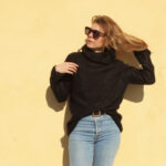 Une femme portant un col roulé chic Pull Mousse col boule taille unique en kid-mohair et soie naturelle et des lunettes de soleil pose contre un mur jaune, ses cheveux flottant dans la brise.