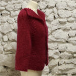 Un mannequin affichant un pull bordeaux en grosse maille contre un mur de pierre blanche texturée.