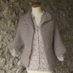 Un cardigan confortable tricoté à la main exposé sur un mannequin contre un mur de pierre texturé, associé à un chemisier à motifs floraux.