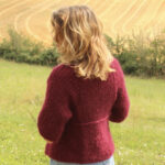 Femme vêtue d'un pull bordeaux debout dans un champ, dos à la caméra, contemplant les collines de la campagne.