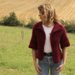 Une personne en tenue décontractée avec un cardigan bordeaux debout dans un champ rural, affichant une expression réfléchie ou pensive.