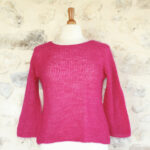 Un Pull Pléïades en mohair de chevreau et soie rose vif exposé sur un mannequin contre un mur de pierre texturée.