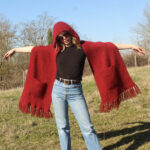 Une personne portant un chapeau rouge et une capuche Etole surdimensionnée assortie en pur mohair de chevreau tricot principal, prenant une pose ludique dans un champ ensoleillé.